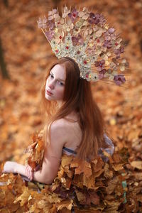 Thoughtful young woman covered with leaves sitting at forest during autumn