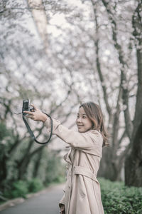 Side view of smiling woman taking selfie with digital camera while standing in park