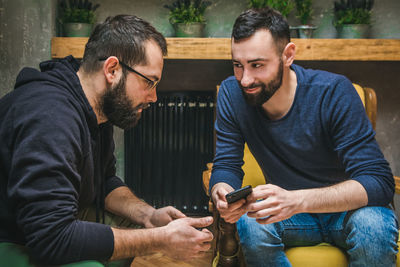 Smiling young man with phone looking at friend sitting at home