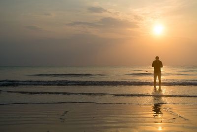 Rear view of silhouette man standing on beach against sky during sunset