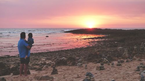 Rear view of father standing with daughter by stacked stones at beach during sunset