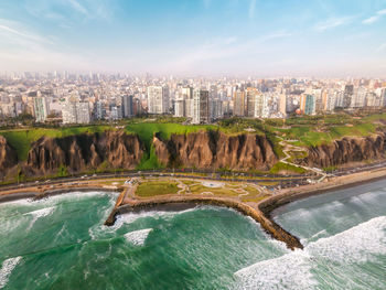 Breathtaking aerial view of wavy ocean washing sandy coastline near modern residential and commercial building against cloudy blue sky in lima