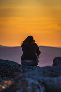 Rear view of teenage girl sitting on rock against sky during sunset