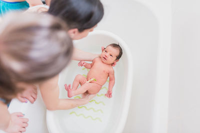 From above unrecognizable parent and kid washing crying newborn baby in warm water in basin at home