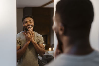 Smiling young man touching face while looking in mirror at home