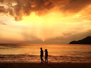 Silhouette people on beach against dramatic sky during sunset