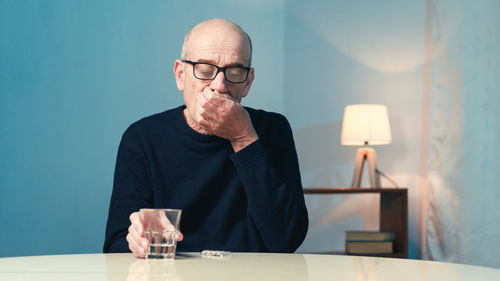 Elderly man with eyeglasses sitting at the table at home eats a heart pill