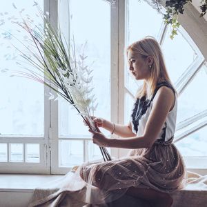 Side view of young woman holding flowers while sitting on window seat