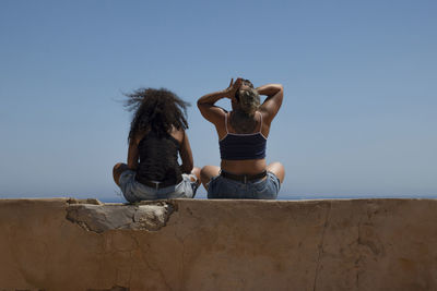 Rear view of 2 women sitting on wall beside the sea against clear blue sky