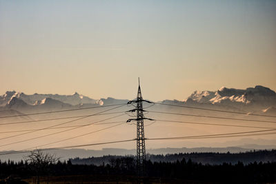 Silhouette electricity pylons by mountains against sky during sunset