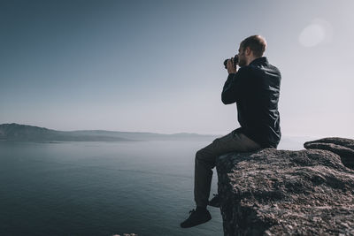 Man sitting on rock looking at sea and mountain with binoculars against sky