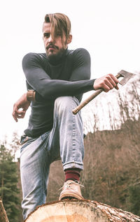 Low angle view of young man with axe standing on log