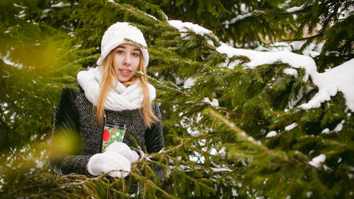 Young woman wearing warm clothing while holding disposable cup amidst trees during winter