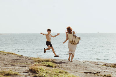 Rear view of woman with bag while son jumping against sea during sunny day