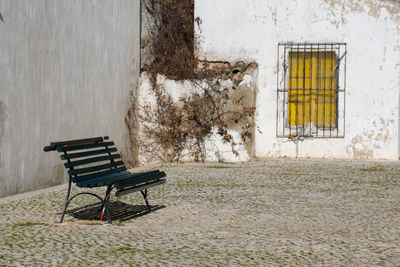 Empty bench against wall with yellow window in front of old building