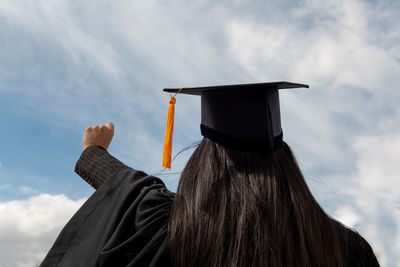 Rear view of woman wearing graduation gown with arms raised standing against cloudy sky