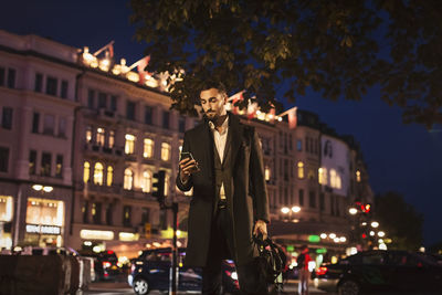 Young man using smart phone while standing on city street at night