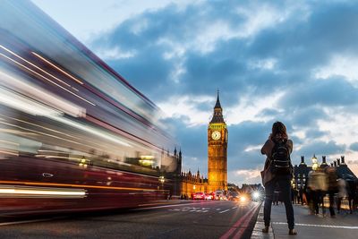 Rear view of woman standing on sidewalk by bus and big ben against cloudy sky during sunset