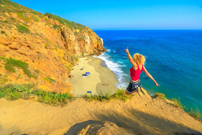 Side view of mature woman with arms raised jumping on cliff by sea against clear blue sky