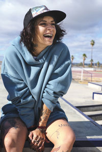 Cheerful female with tattoos and in stylish outfit sitting on street in city and laughing at joke