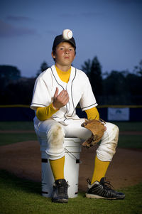Portrait of boy in baseball uniform sitting on bucket tossing baseball