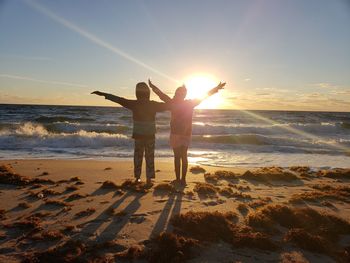 Rear view of people standing at beach against sky during sunset