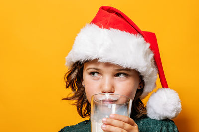 Cute girl in santa hat sipping milk from glass and looking away during christmas celebration against yellow background