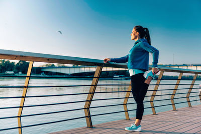 Young woman exercising on promenade by river against clear sky during sunset