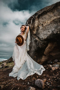 Woman wearing white dress while leaning on rock at beach against cloudy sky