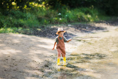 Preschooler boy in yellow rubber boots plays fishing, child fishes with stick  in countryside