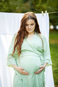White pretty pregnant woman stands putting hands on big belly. green trees, park,white sheet on