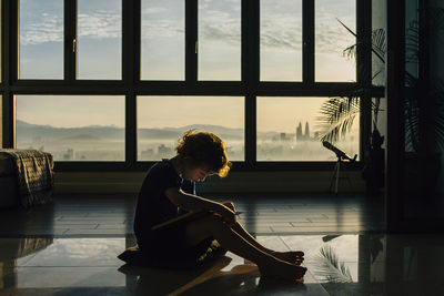 Full length of boy reading book while sitting on floor against windows at home