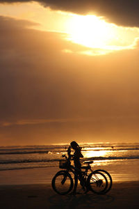 Silhouette riding bicycle on beach against sky during sunset