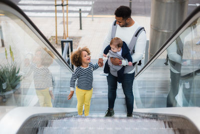 From above of positive young ethnic man carrying infant son and holding hand of cheerful little daughter while standing on escalator in city