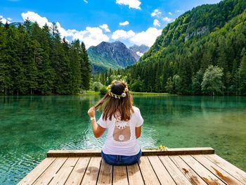 Rear view of woman sitting on pier at lake against mountains