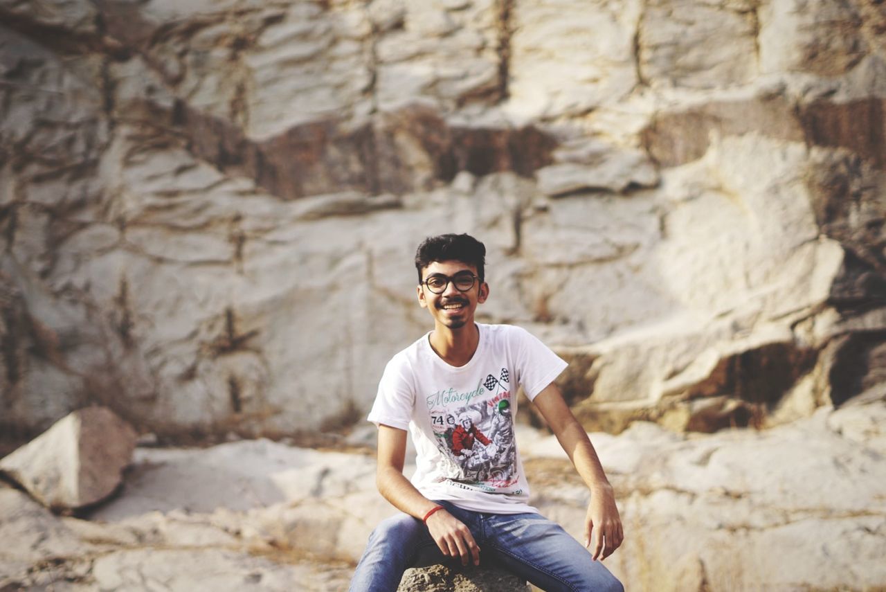 young men, real people, three quarter length, leisure activity, lifestyles, young adult, one person, sunglasses, casual clothing, fashion, portrait, glasses, front view, rock, sitting, looking at camera, rock - object, solid, focus on foreground, outdoors