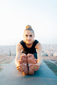 Full body of flexible female practicing paschimottanasana on sports mat on rooftop at sunset