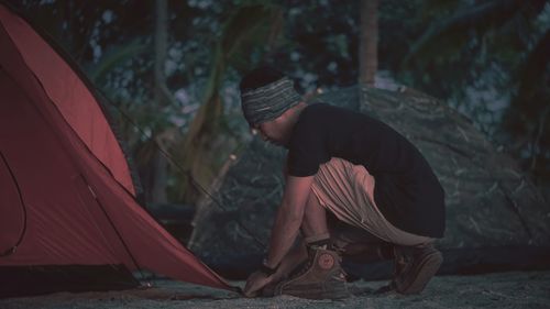 Side view of man placing tent in forest at night