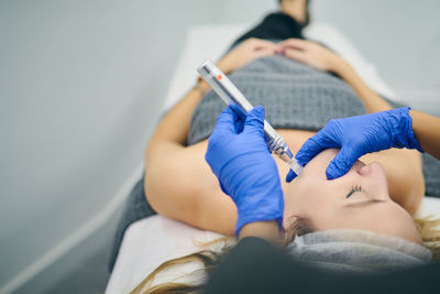 Cropped unrecognizable cosmetologist in uniform using derma pen with micro needles and cleaning face of client in beauty clinic