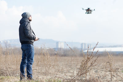 Side view of mid adult man flying drone while standing on field against sky