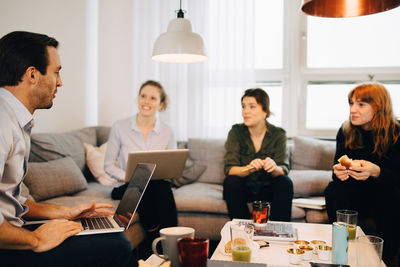 Female professionals looking at businessman sitting with laptop on sofa in creative office