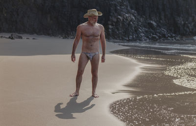 Full length of shirtless man with swinwear and cowboy hat standing on beach