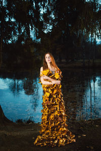 Thoughtful young woman covered with leaves standing by lake at forest during autumn