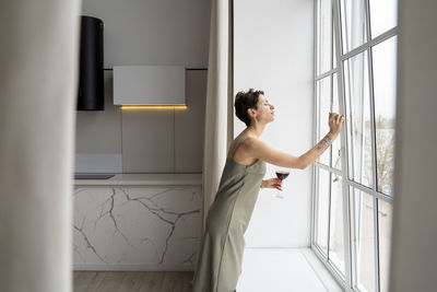 Woman holding wineglass opening window at home