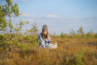 Stylish young woman in hipster hat and coat gathers cranberries in meadow grass