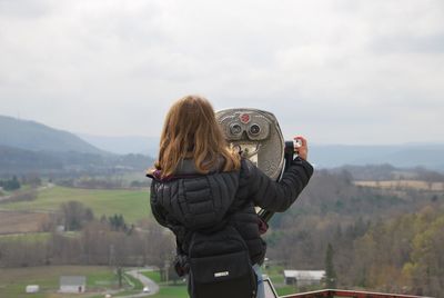 Rear view of woman standing with coin-operated binoculars against sky