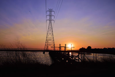 Silhouette electricity pylon by lake against sky during sunset