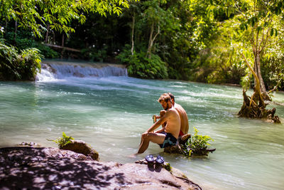 Full length of shirtless man sitting at riverbank in forest