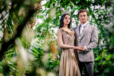 Portrait of confident bride and groom holding hands while standing amidst plants