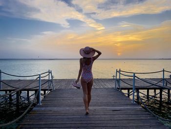 Woman wearing swimwear and summer hat walking down a wooden pontoon towards the sea at sunset